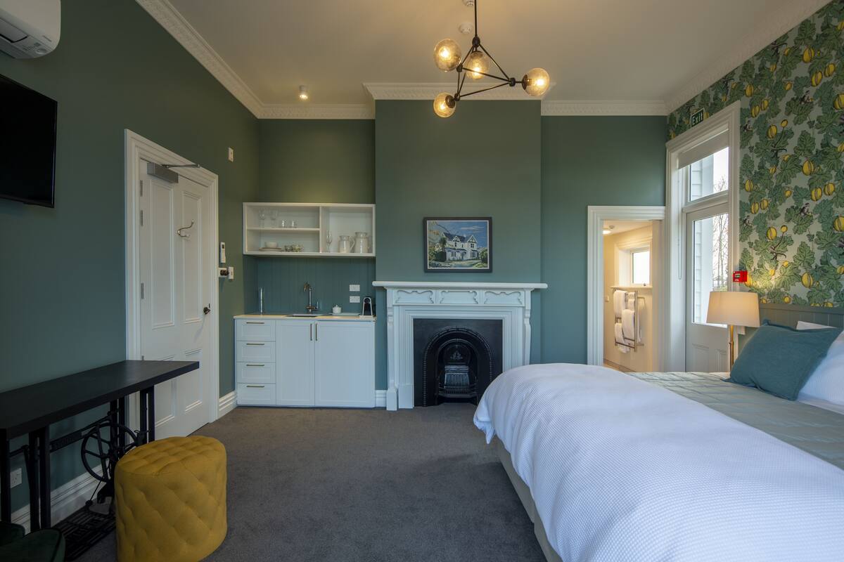 A bedroom with a bed, a fireplace, a desk, and a yellow ottoman.