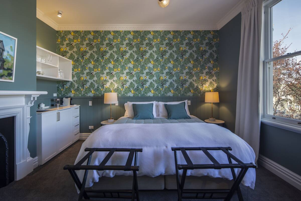 A bedroom with a large bed, two bedside tables, a nightstand, a dresser, a fireplace, and a window with a view.
