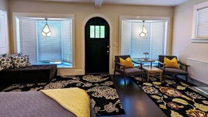 Una habitación con una cama, un escritorio, dos sillas, una puerta negra y ventanas con persianas.
