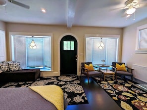 A room with a bed, a desk, two chairs, a black door, and windows with blinds.