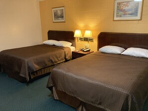 A hotel room with two beds, each with a pillow and a blanket. There is a nightstand with a lamp and a phone.