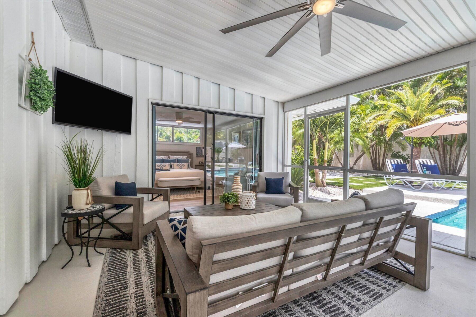 A modern living room with a sofa, a coffee table, and a TV. There is a sliding glass door leading to a pool area with lounge chairs.