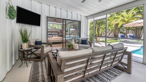 A modern living room with a sofa, a coffee table, and a TV. There is a sliding glass door leading to a pool area with lounge chairs. - Newly remodeled beach house by Lido Beach and private pool (Sarasota)