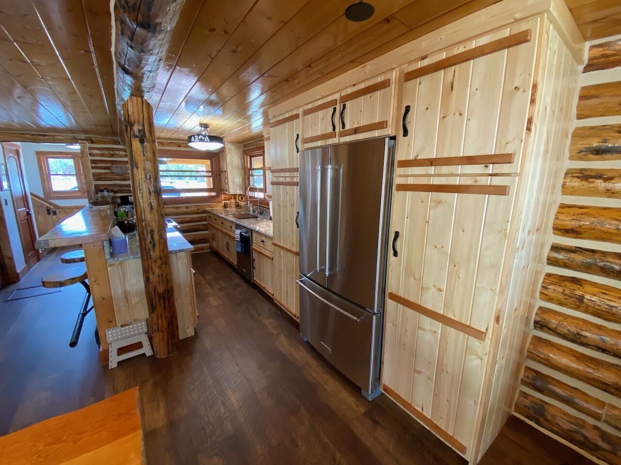 A wooden cabin interior with a kitchen featuring a large stainless steel refrigerator, wooden cabinets, and a dining area with a bench.