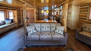 Ein Interieur einer Blockhütte mit einer zentralen Sitzgruppe, bestehend aus einer Couch und Sesseln, Holzböden und einer Küche im Hintergrund.