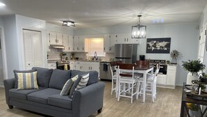 A modern living room with a grey sofa, dining table, and white chairs. There is a kitchen area with white cabinets and a stainless steel refrigerator.