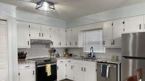 A modern kitchen with white cabinets, a stainless steel refrigerator, a built-in oven, and a window with blinds.