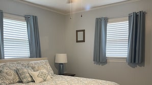 A bedroom with a bed, a ceiling fan, a nightstand with a lamp, and two windows with curtains.