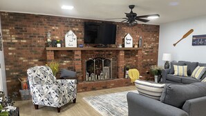 A living room with a brick fireplace, a sofa, a patterned armchair, and a television mounted above the fireplace.