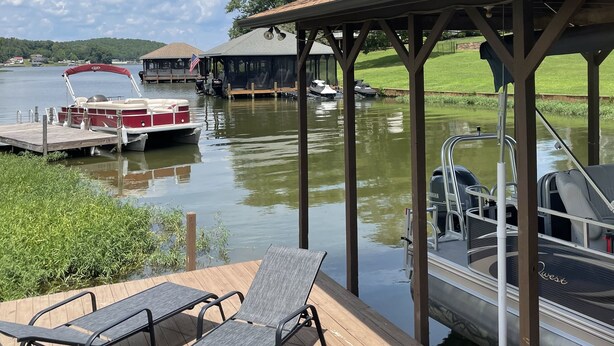 A wooden deck with lounge chairs, a covered area with a roof, and boats docked at a calm water body.