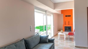 A modern living room with a grey sofa, a white coffee table, and a red chair. There is a fireplace with a TV above it, and a sliding glass door leading to an outdoor area. - Athens & Acropolis View 7th Floor Apartment (Athens)
