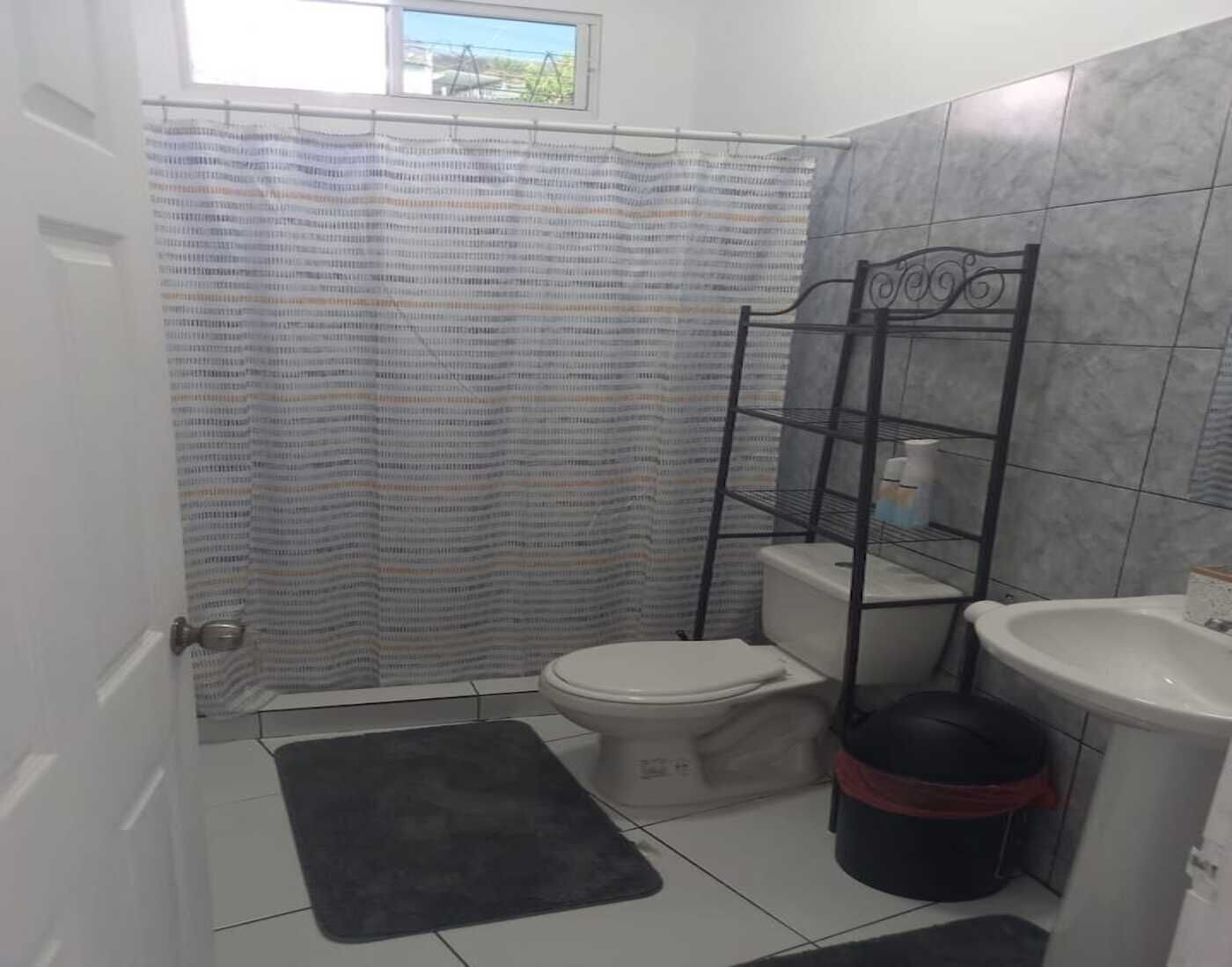 Un baño con inodoro, lavamanos, un cubo de basura negro, una estantería metálica y una cortina de ducha estampada.