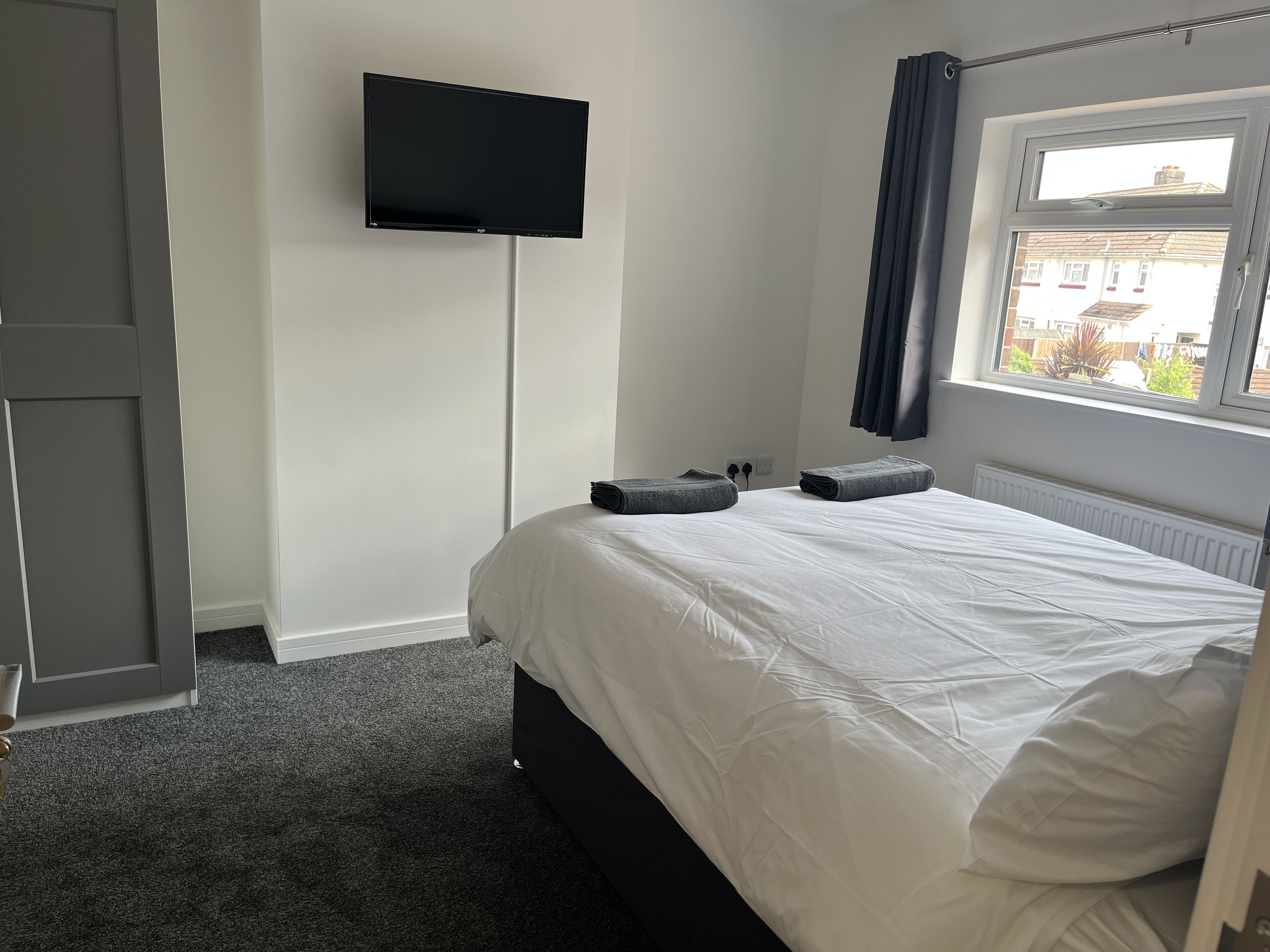A bedroom with a bed, a flat-screen TV mounted on the wall, a window with curtains, and a radiator.