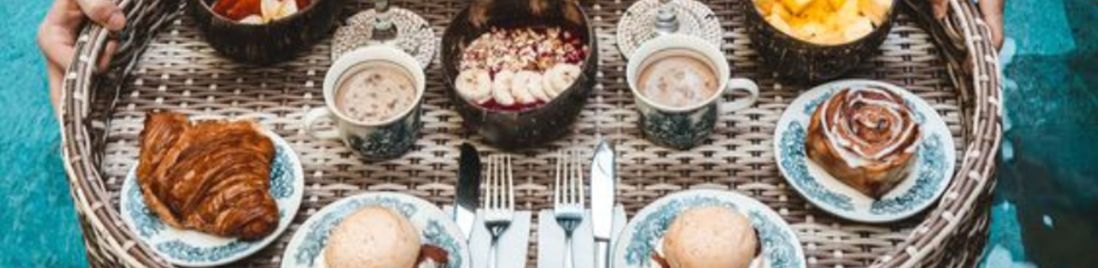 A wicker tray with various food items and drinks, including pastries, a croissant, and a bowl of soup.