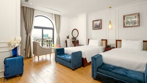 Habitación de hotel con dos camas, una zona de estar, un ventanal grande con cortinas, una maleta y vistas a la ciudad.