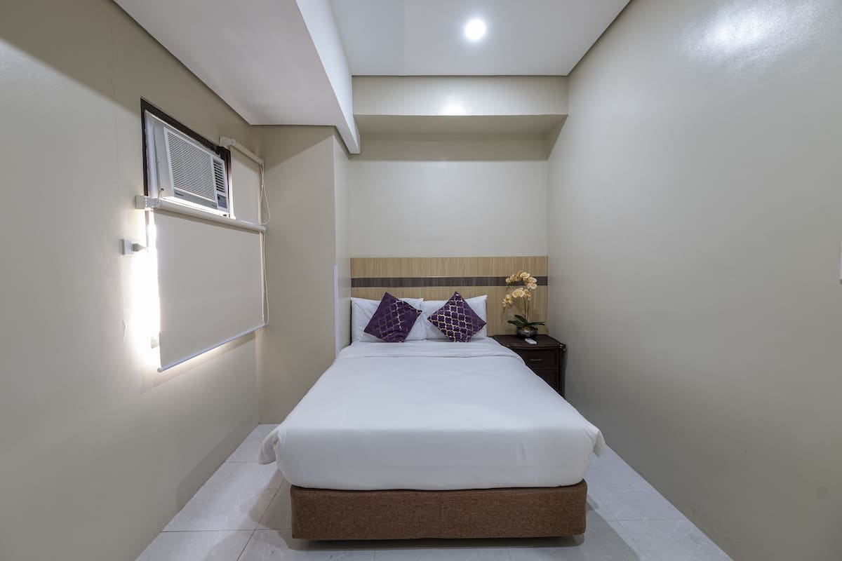 Une chambre d’hôtel bien rangée, avec un lit, une table de chevet, un climatiseur fixé au mur et un luminaire au plafond.