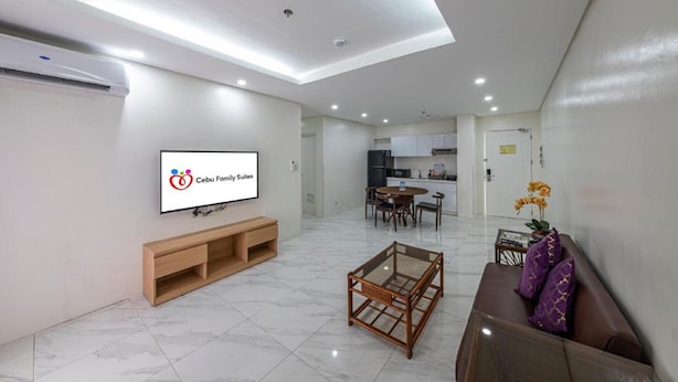 A modern living room with a flat-screen TV, a wooden entertainment unit, a glass-top coffee table, a brown sofa with purple cushions, and a dining area with a table and chairs. - Cebu Family Suites powered by Cocotel (Cebu City)