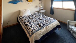 Une chambre à coucher comprenant un lit, un ventilateur de plafond, une fenêtre avec des rideaux, une petite table avec une lampe et des éléments de décoration murale, notamment un poisson et un anneau de sauvetage.