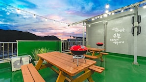 A rooftop patio with a barbecue grill, picnic tables, and string lights against a sunset sky.