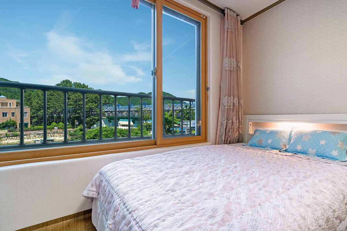 Un dormitorio con un ventanal grande, una cama con colcha estampada y vistas a un parque y edificios.