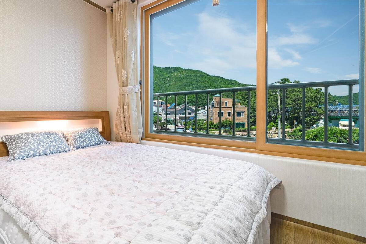 Un dormitorio con un ventanal grande, una cama con ropa de cama floral y vistas a un paisaje verde y edificios.