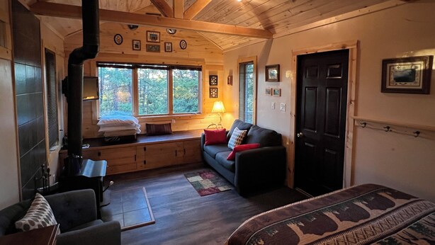 Hyggelig hyttekrog med pejs, træbjælker, sofa og vindue med udsigt til træer.