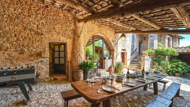 A rustic stone interior with a long dining table set for a meal, surrounded by potted plants and a foosball table.