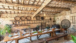 A rustic dining area with wooden tables and benches, stone walls, and a thatched roof.