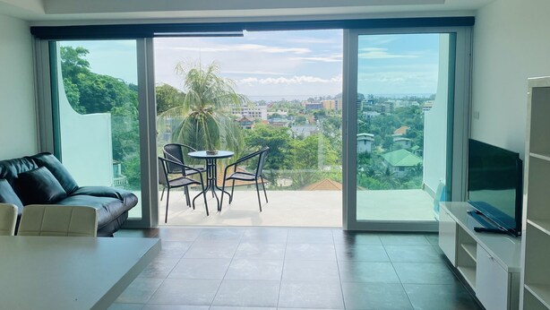 A modern living room with a large sliding glass door leading to a balcony, a sofa, a TV, and a dining area with chairs. - Sea view apartment, 5 min drive from Kata Beach (Karon)