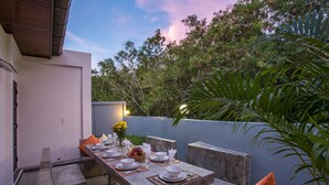Un área de comedor al aire libre con una mesa dispuesta para cuatro personas, rodeada de escalones y bancos de piedra, con vista a árboles y un cielo despejado.