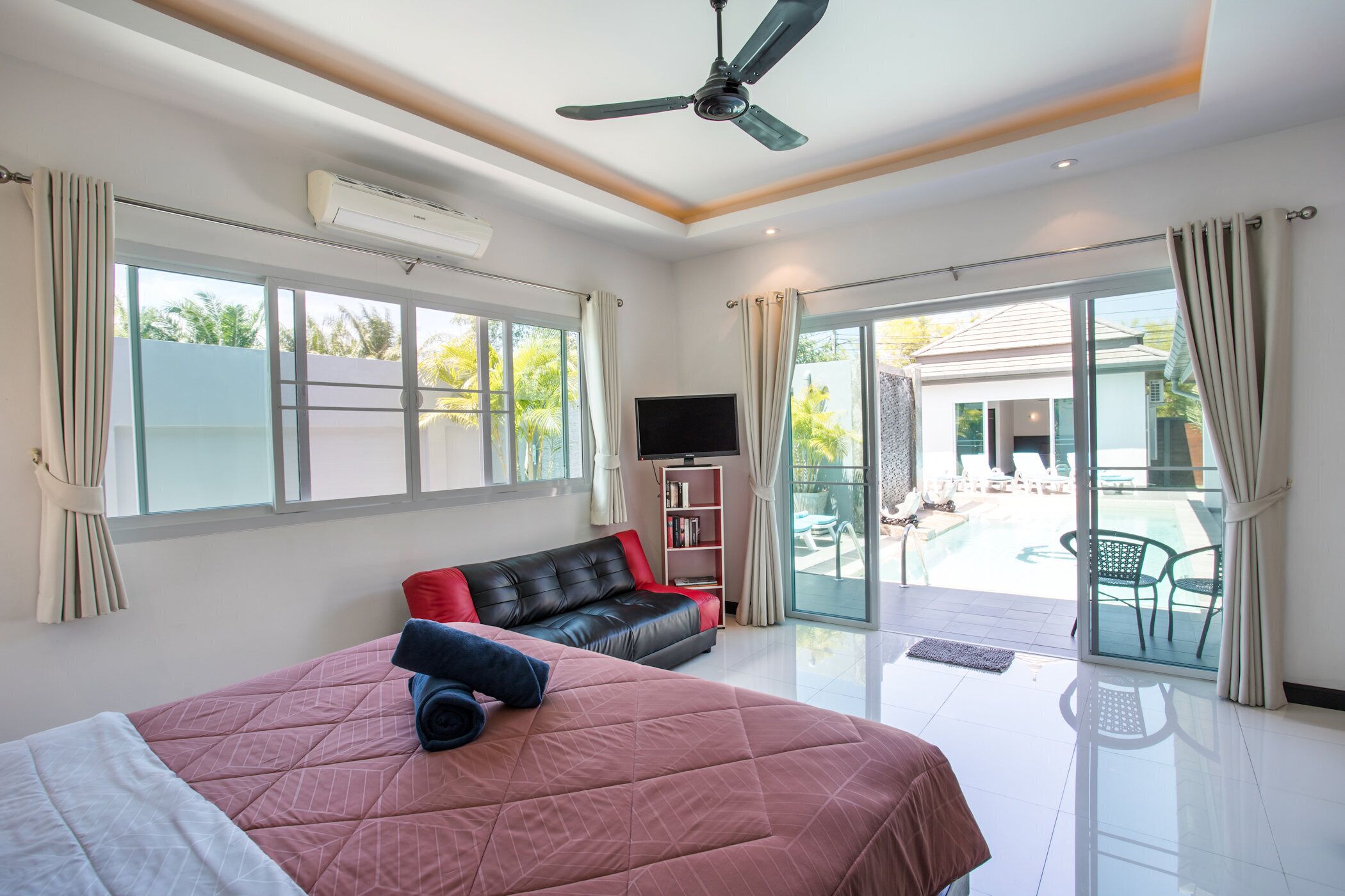 A spacious bedroom with a large bed, a ceiling fan, a TV, and a sliding glass door leading to a pool area.