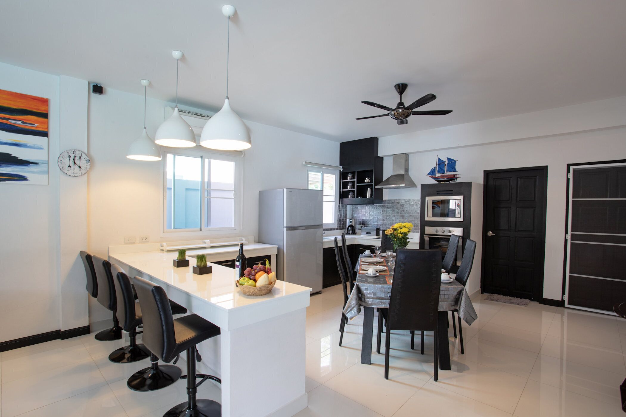 A modern kitchen with a central island, bar stools, a dining table with chairs, a refrigerator, an oven, and a ceiling fan.