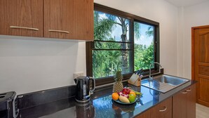 A kitchen with wooden cabinets, a black countertop, a window with a view of palm trees, a sink, and a fruit bowl with various fruits. - Tropical Vibe 5BR Pool Villa Nikkie 3 (Rawai)