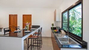 A modern kitchen with a large window, a dining area with chairs, and a wooden door. - Tropical Vibe 5BR Pool Villa Nikkie 3 (Rawai)
