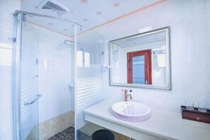 Un baño con cabina de ducha de vidrio, un lavamanos blanco con diseño morado, un espejo con marco decorativo y un pequeño armario de madera con vasos.