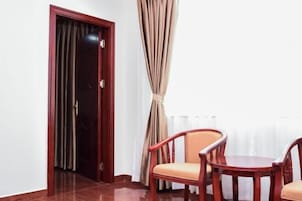 Una habitación con una puerta de madera, una ventana con cortinas color beige y dos sillas de madera alrededor de una mesa pequeña.