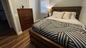 A bedroom with a wooden bed, a dresser, a nightstand, and a window with blinds. - The Nantucket Suites  (Elizabethtown)