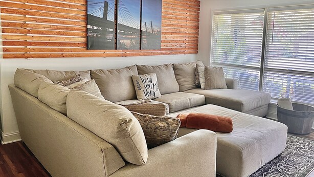 A living room with a sectional sofa, a ceiling fan, and a large window with blinds. - The Nantucket Suites  (Elizabethtown)
