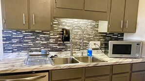 A kitchen with a stainless steel sink, a dish rack, a microwave, and a tiled backsplash. - The Nantucket Suites  (Elizabethtown)