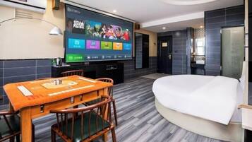 A hotel room with a flat-screen TV mounted on the wall, a wooden dining table with chairs, a bed with a white mattress, and dark tiled flooring.