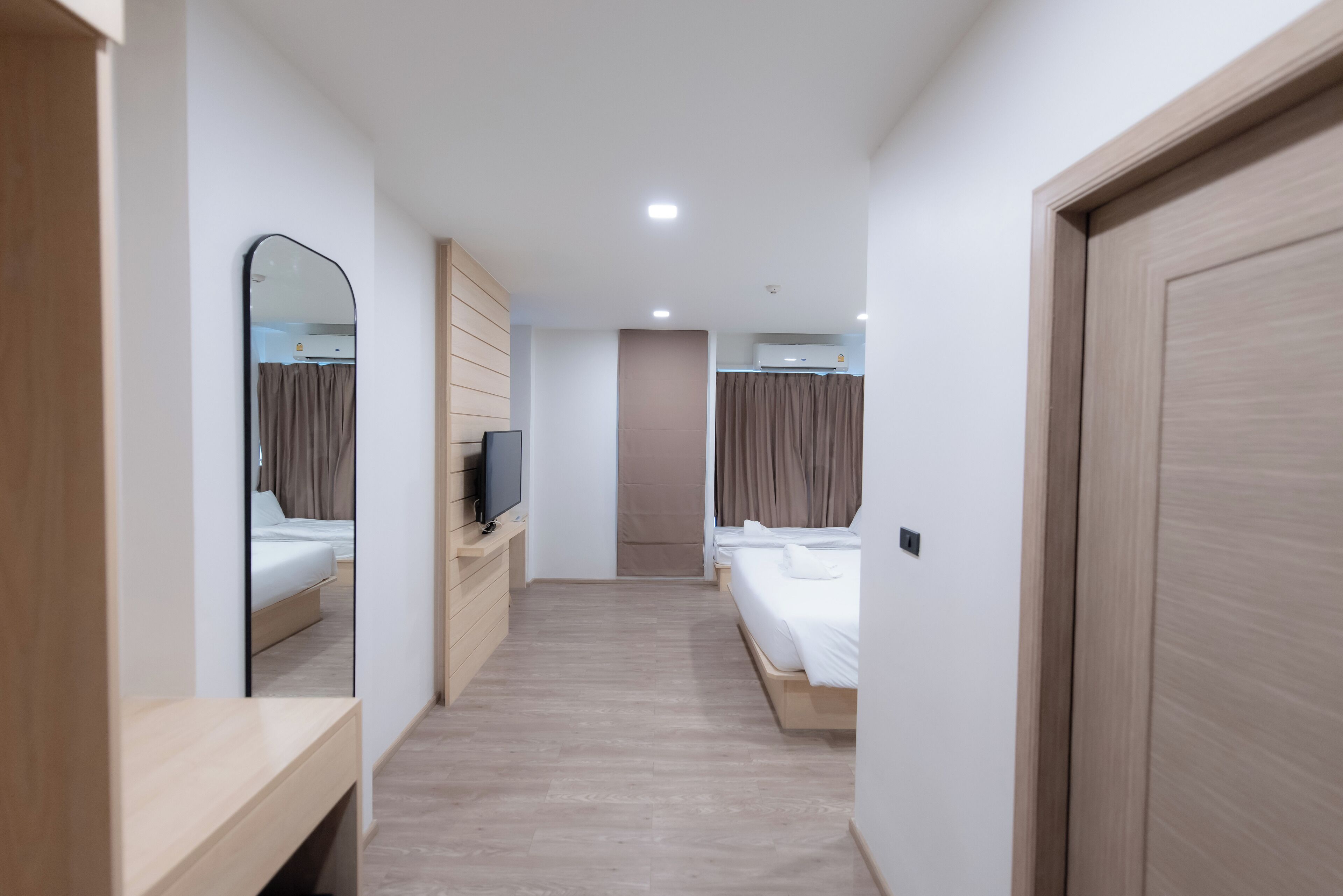 A hotel room with a wooden floor, a large mirror, a TV on a wooden stand, two beds with white linens, and a door with a light switch.