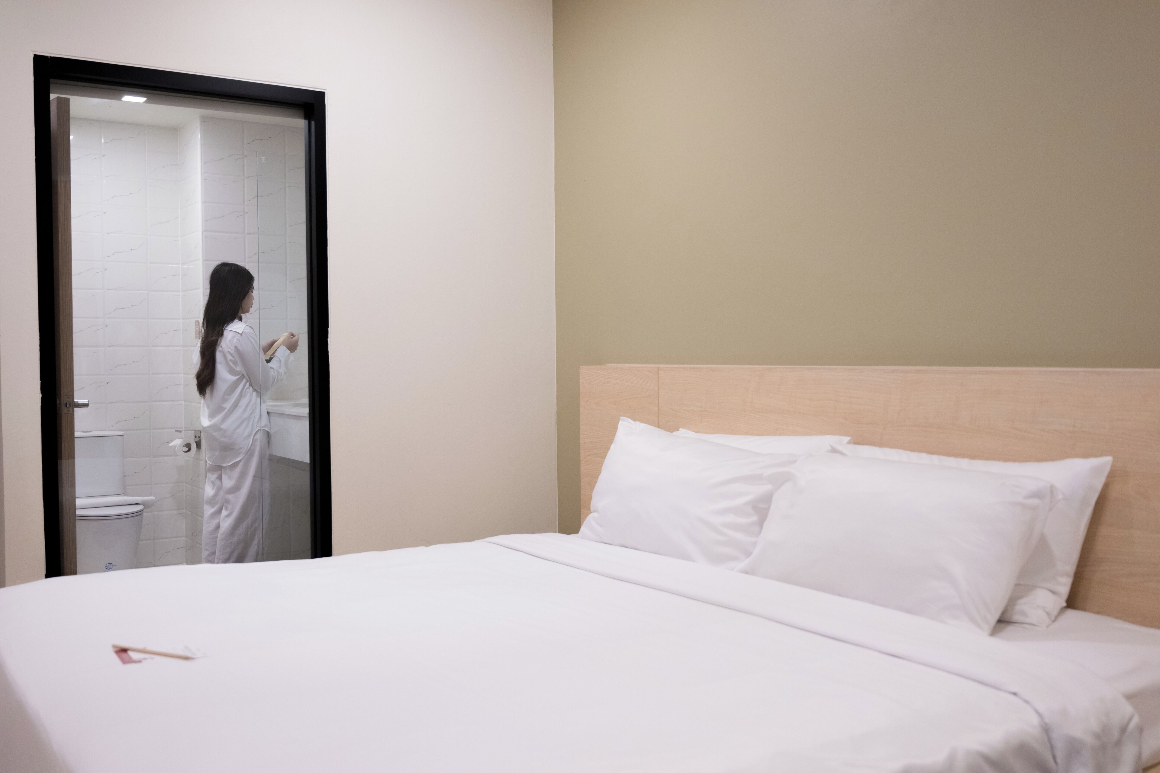 A hotel room with a bed, white linens, a wooden headboard, and a bathroom visible through a glass door.