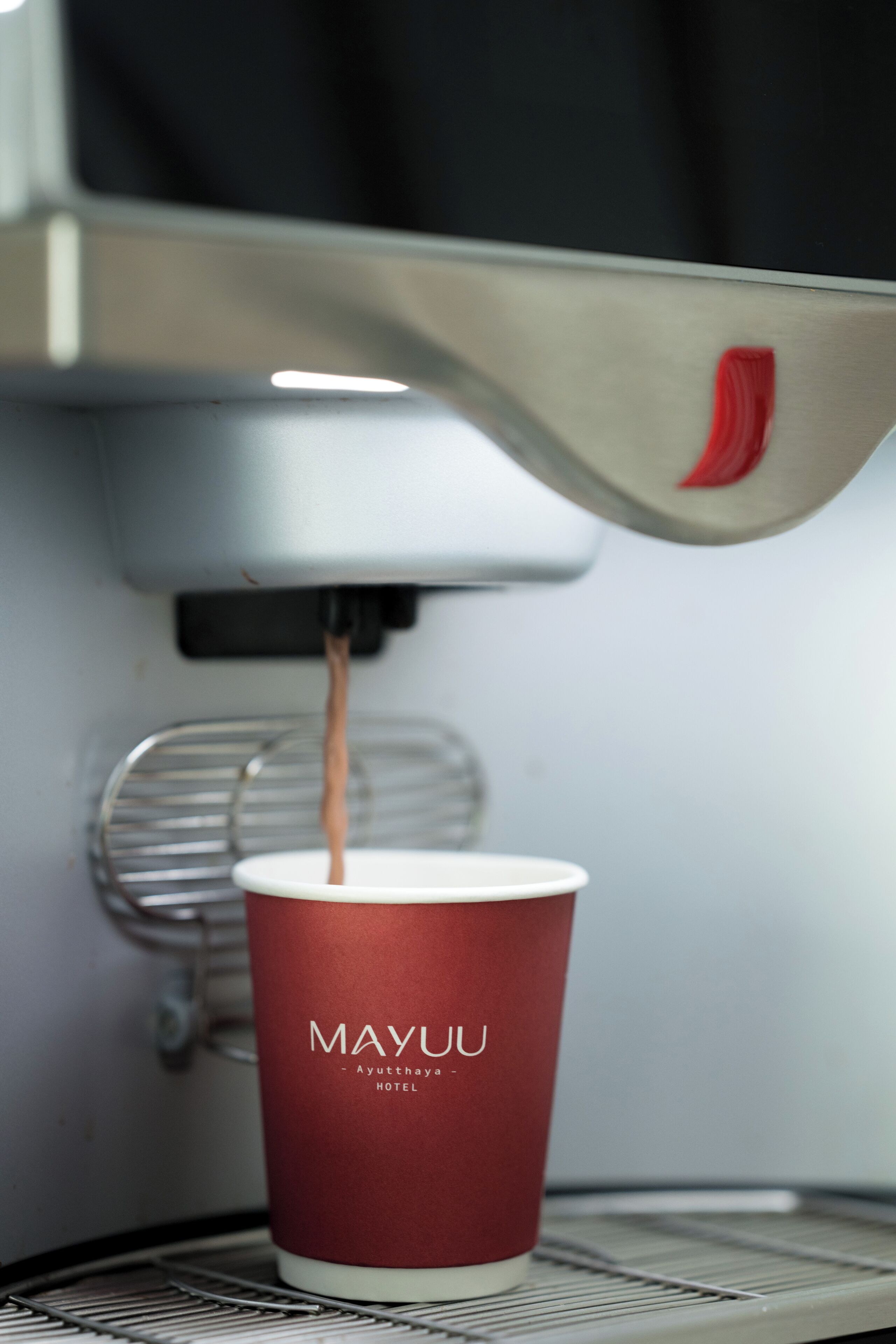 Un gobelet en papier rouge, portant l’inscription « MAYUU – Hôtel d’Ayutthaya », est rempli d’une boisson provenant d’un distributeur automatique.