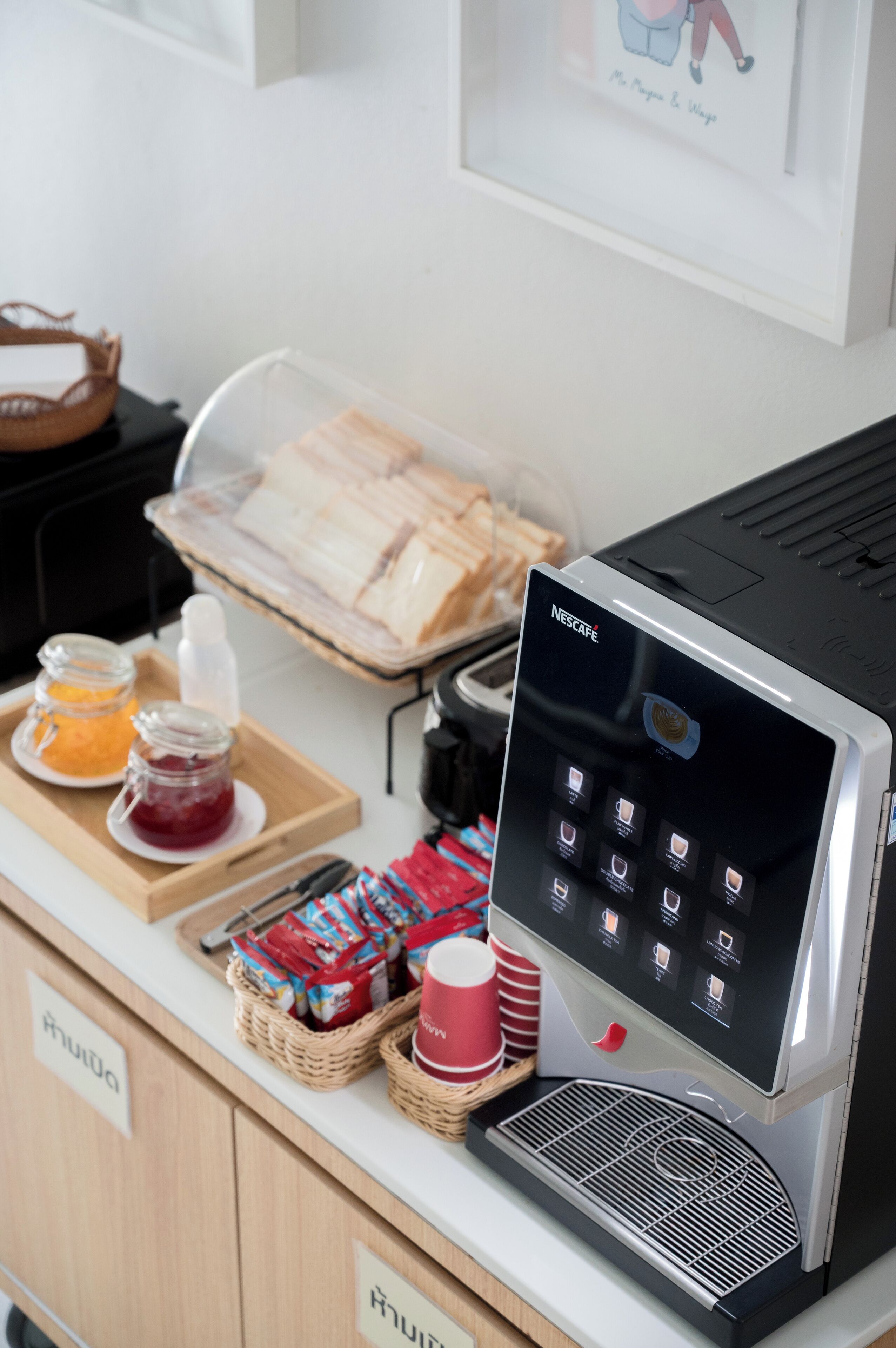 A Nescafe coffee machine with a touchscreen interface, a tray of bread, and a selection of condiments and beverages on a counter.