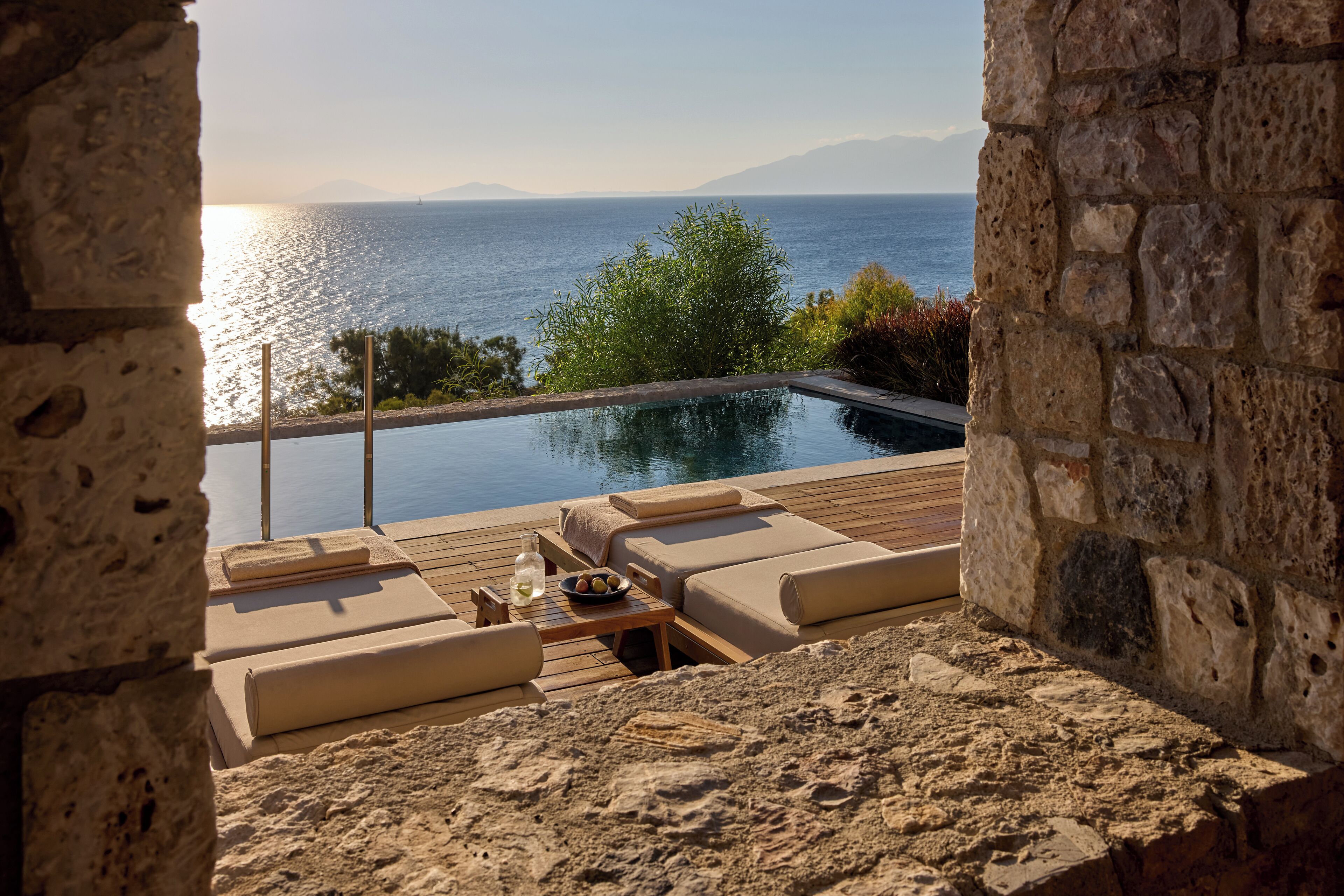 A stone-walled infinity pool with beige loungers and a wooden deck, overlooking a calm sea and distant mountains.
