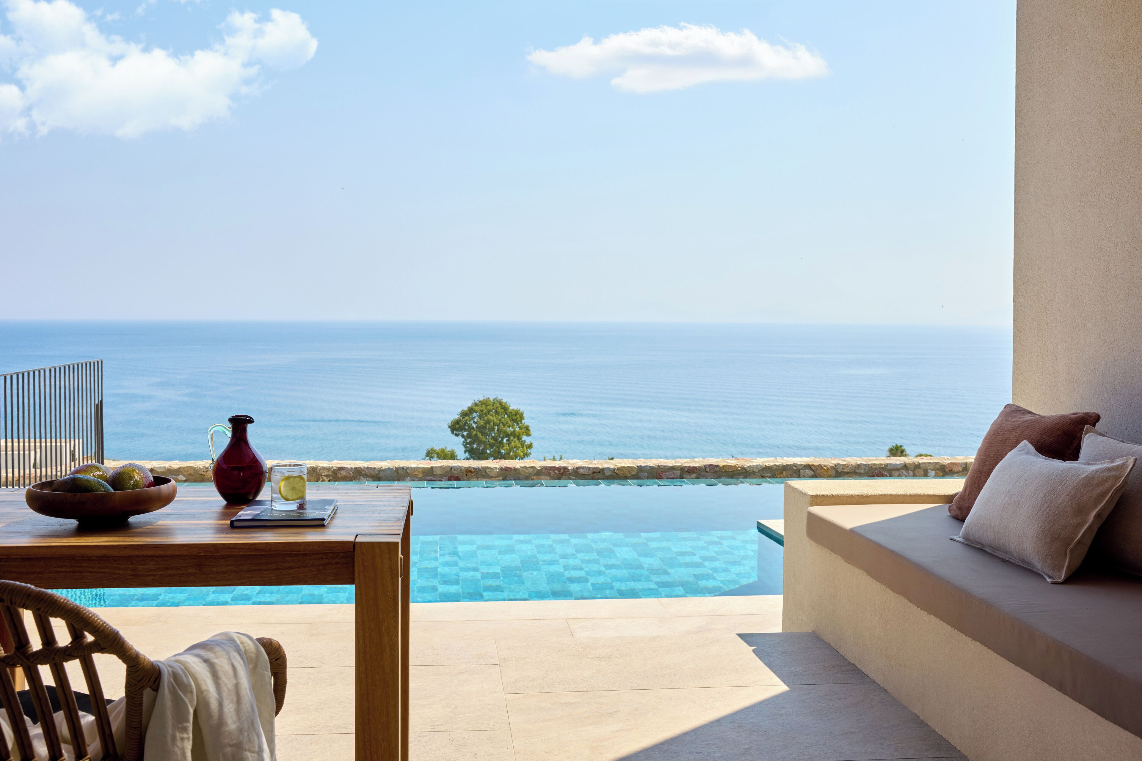 Un balcon avec vue sur la mer, une table avec un saladier de fruits, une carafe et un verre rempli de boisson.