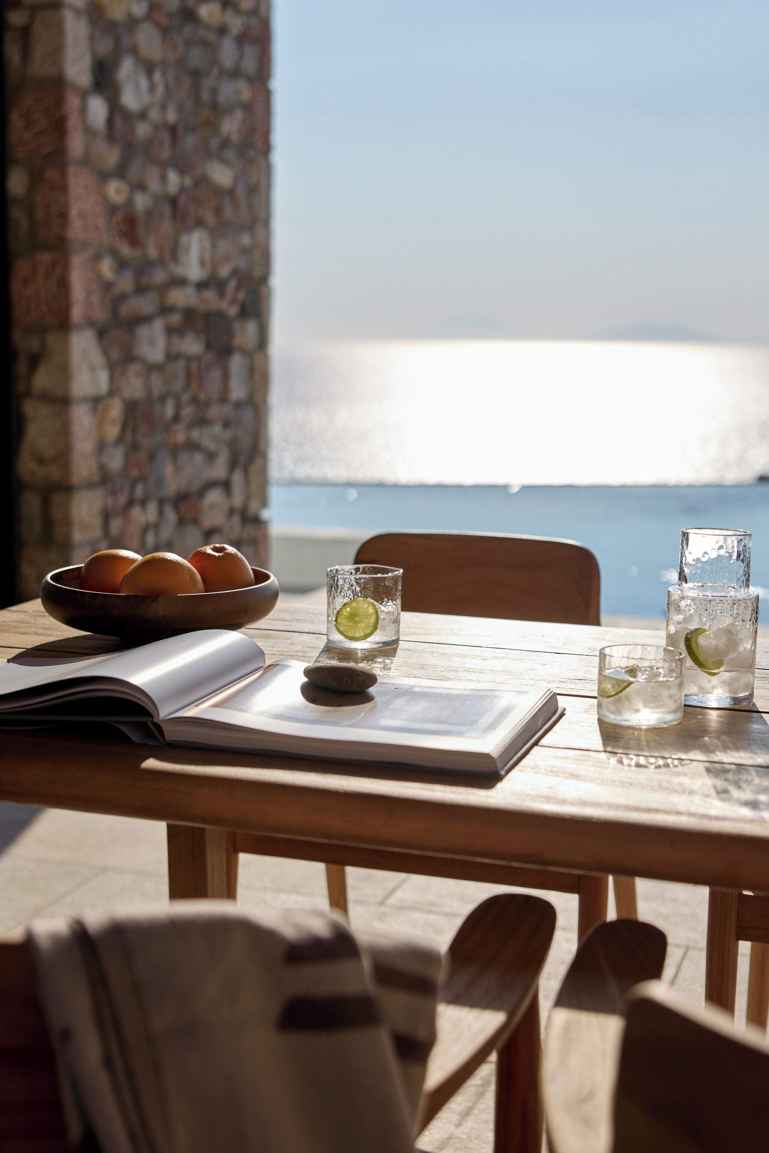 Une table en bois sur laquelle se trouve un saladier rempli d’oranges, deux verres d’eau avec des quartiers de citron vert et un livre.
