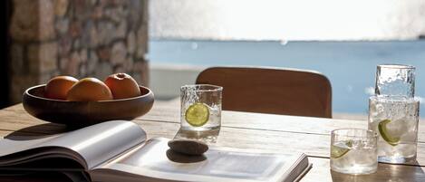 Une table en bois sur laquelle se trouve un saladier rempli d’oranges, deux verres d’eau avec des quartiers de citron vert et un livre.