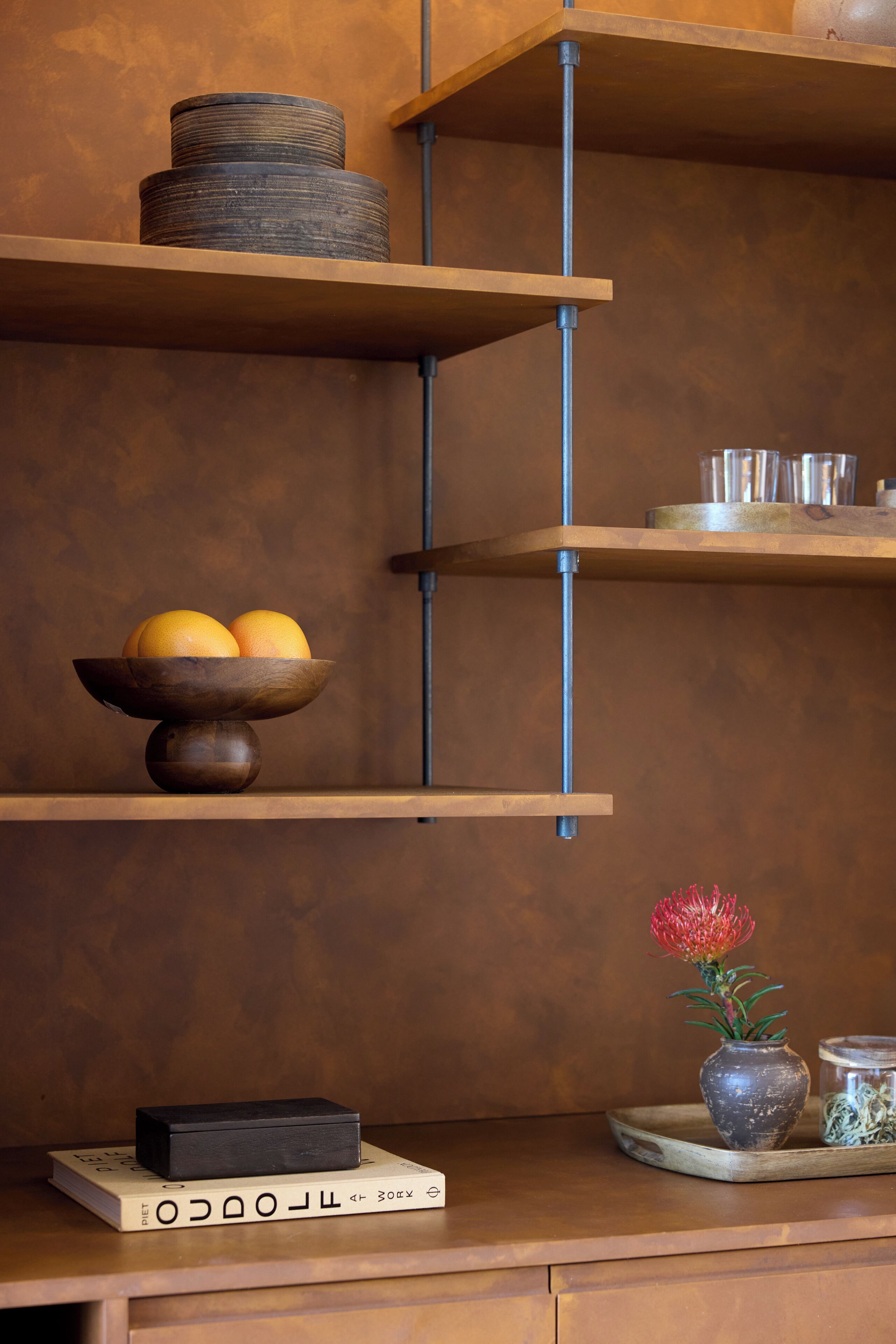 Ein hölzernes Regal mit einer Schale Orangen, einer Vase mit einer roten Blume und einem Buch mit dem Titel "The Obstacle".