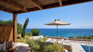 A poolside area with a view of the sea, a table with a bowl of fruit, and a lounge chair under an umbrella. - KOIA All - Suite Well Being Resort - Adults Only (Kos)